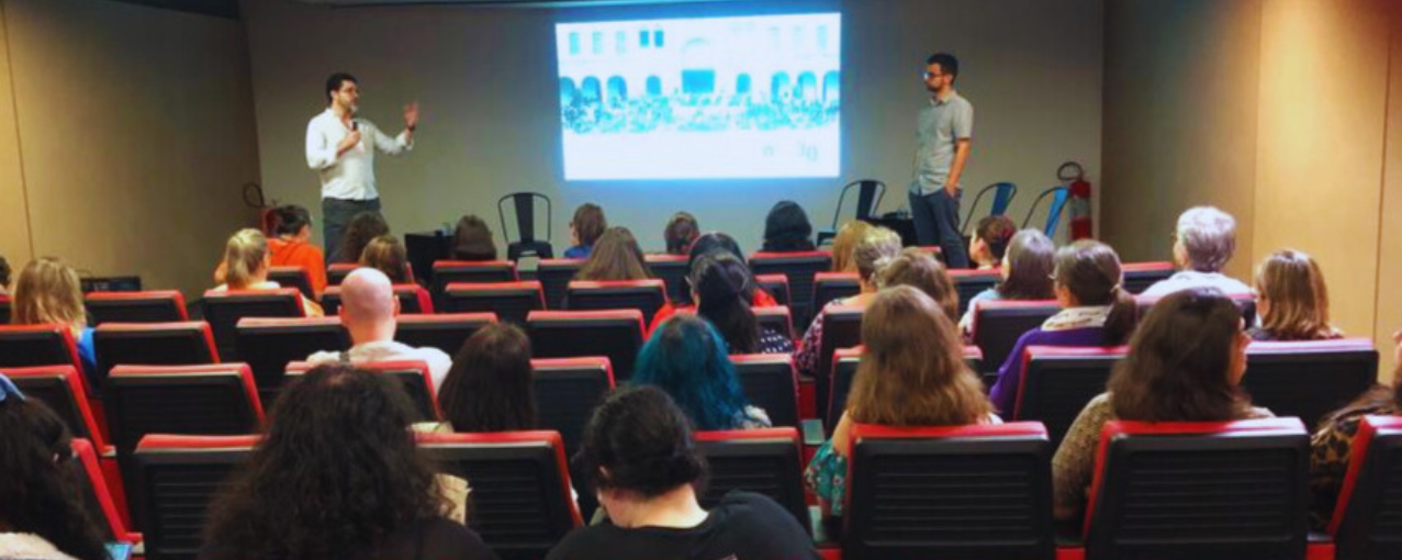 Fotografia do auditório, duas pessoas no palco, em frente ao telão e público sentado na plateia.